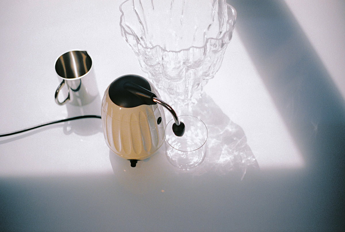 Cream Mokapod electric moka maker beside a glass cup on a sunlit table
